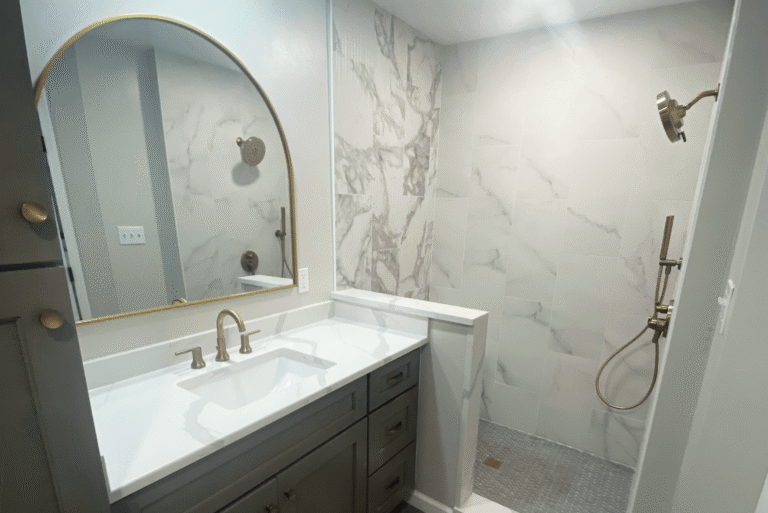 Elegant bathroom remodel featuring a large walk-in shower with marble-look tile, dual rain shower heads in brushed gold, a matching gold faucet, and a white vanity with quartz countertop and arched gold-framed mirror.