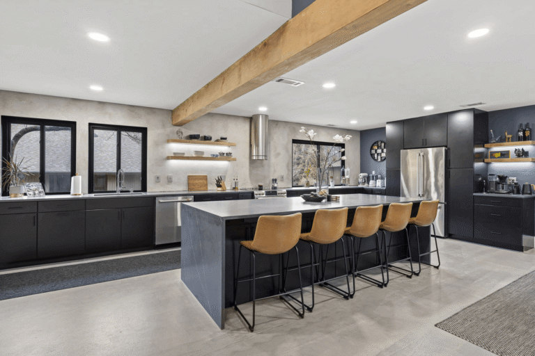 Contemporary kitchen remodel in Northern Virginia by Veteran Built Homes featuring flat-panel black cabinetry, oversized waterfall island with gray quartz countertop, leather bar stools, floating wood shelves, stainless steel appliances, and exposed wood beam for a modern industrial design.