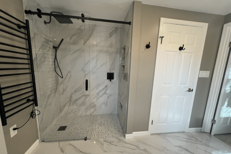 Master bathroom remodel in Springfield Virginia featuring frameless sliding glass shower enclosure with large-format Calacatta marble-look tile, matte black rainfall shower head and fixtures, basketweave mosaic tile shower floor, matching marble-look floor tile, and matte black heated towel rack by Veteran Built Homes