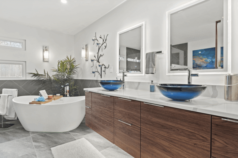 Luxury master bathroom remodel in Northern Virginia featuring floating walnut double vanity with white quartz countertop, dual cobalt blue glass vessel sinks, backlit LED mirrors, freestanding oval soaking tub, large-format slate tile flooring, and designer wall sconces by Veteran Built Homes