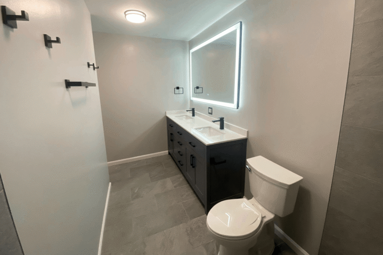 Modern bathroom remodel in Northern Virginia by Veteran Built Homes featuring a dark double-sink vanity with matte black fixtures, LED backlit mirror, minimalist towel hooks, and sleek gray tile flooring for a clean and contemporary design.
