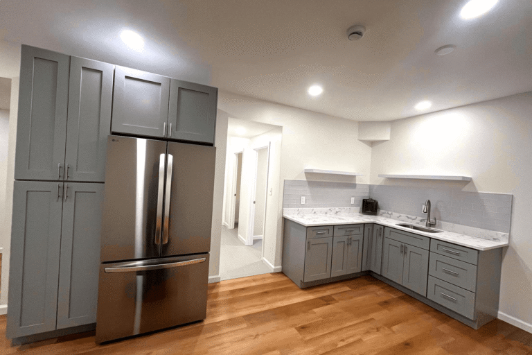 Modern basement kitchen remodel in Northern Virginia by Veteran Built Homes featuring grey shaker cabinets, stainless steel refrigerator, marble countertops, subway tile backsplash, and warm wood flooring.
