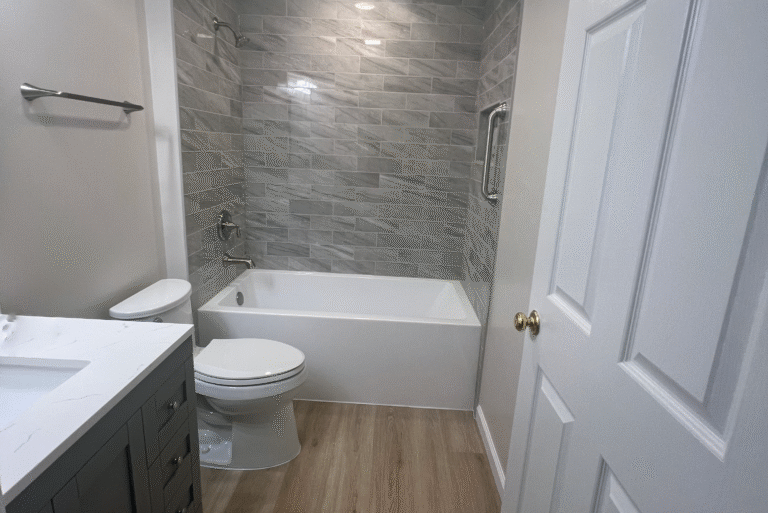 Modern bathroom remodel featuring gray subway tile shower surround, white bathtub, white toilet, gray vanity with marble countertop, and wood-look vinyl flooring.