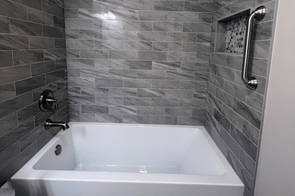 Close-up of a modern bathtub with gray marble-look tile surround, built-in niche with mosaic tile accent, and stainless steel grab bar for safety.