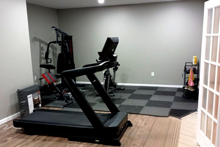 Finished basement home gym remodel in Northern Virginia by Veteran Built Homes featuring treadmill, weight training equipment, padded flooring, and bright recessed lighting.
