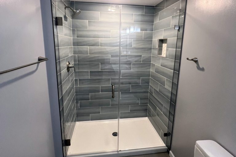 Basement bathroom upgrade in Fairfax Virginia featuring oversized frameless glass walk-in shower with floor-to-ceiling gray stacked tile, rainfall shower head, handheld sprayer, and recessed lighting by Veteran Built Homes