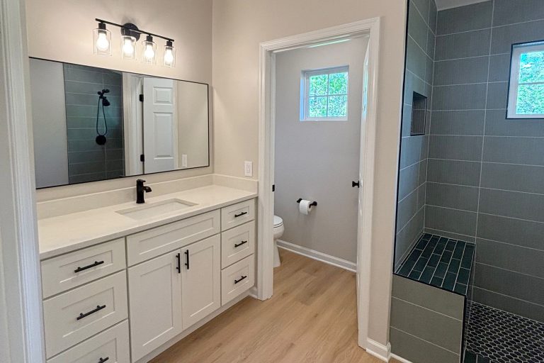 "Farmhouse modern bathroom remodel in Bristow Virginia featuring white shaker vanity with quartz countertop, matte black fixtures, industrial vanity lighting, walk-in shower with gray tile and hexagon floor tile, and wood-look vinyl flooring by Veteran Built Homes
