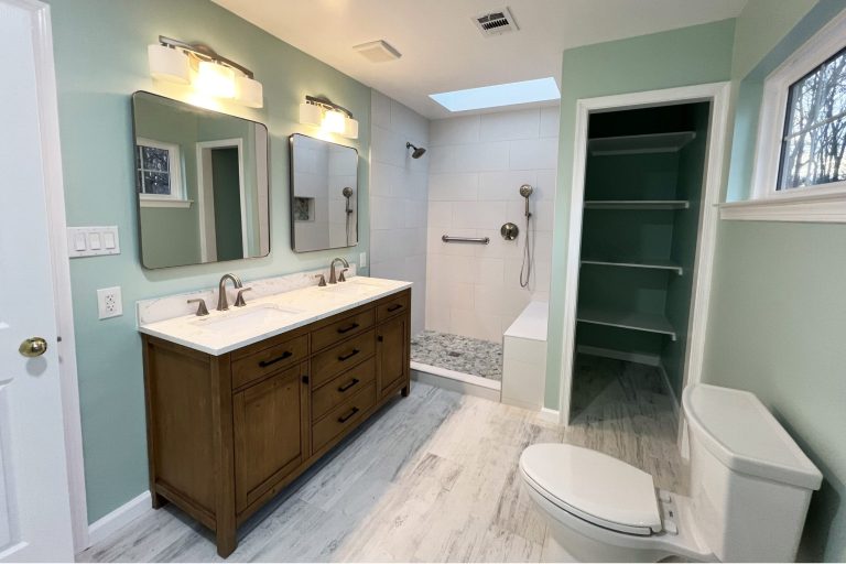 Bathroom remodel in Herndon Virginia featuring walk-in shower with white subway tile, skylight, pebble tile floor, brushed nickel grab bar and handheld sprayer, double rustic wood vanity with quartz countertop, wood-look tile flooring, and built-in open shelving by Veteran Built Homes