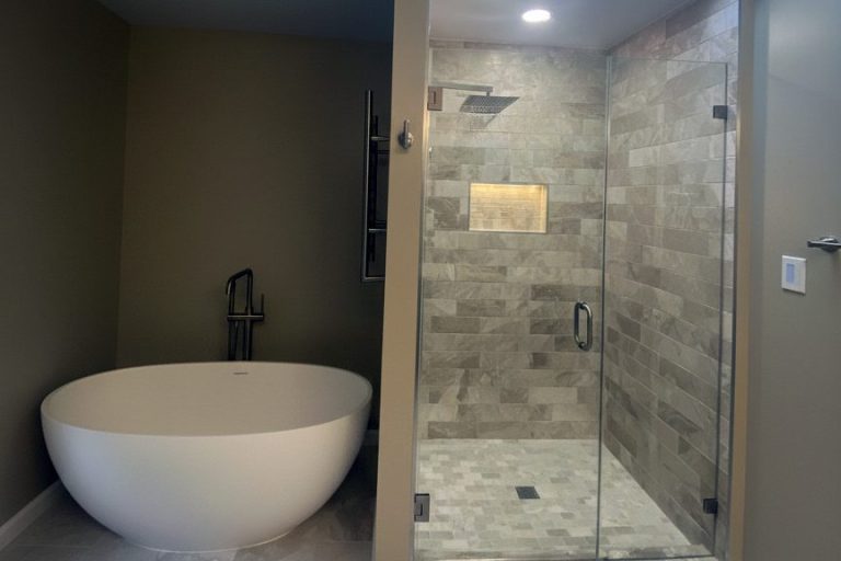 Full master bathroom remodel in Springfield Virginia featuring freestanding oval soaking tub with floor-mount faucet, frameless glass walk-in shower with large-format gray stone tile, rainfall shower head, and illuminated built-in niche by Veteran Built Homes