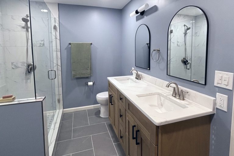 Modern bathroom remodel in Montclair Virginia featuring frameless glass walk-in shower with marble-look porcelain tile, double sink vanity with quartz countertop, matte black hardware, dual arched mirrors, and slate tile flooring by Veteran Built Homes