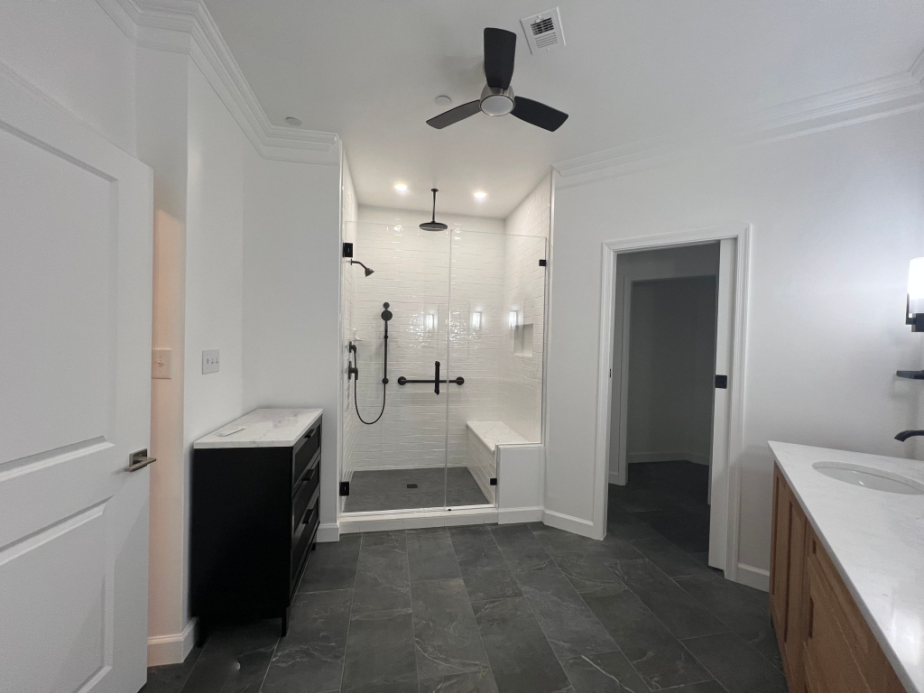 A wide-angle view of a spacious, modern bathroom featuring dark gray tile floors, crisp white walls with crown molding, and a central frameless glass walk-in shower. The shower includes white subway tile, a built-in bench, and matte black fixtures. A black cabinet with a marble top sits to the left, and a light wood vanity is partially visible on the right.